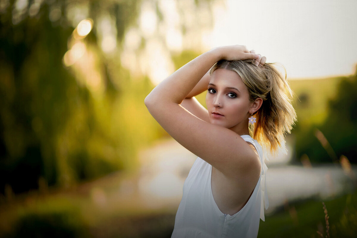 A woman in a white top holds her hair up while standing in a park at sunset for her Pella Senior Photographer