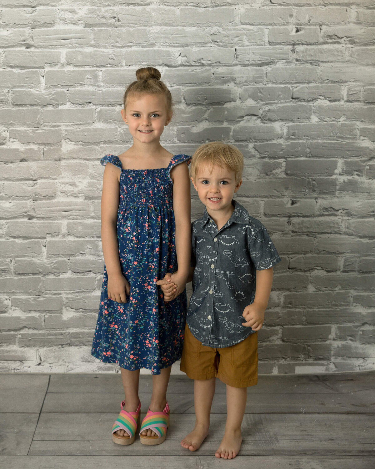 Big sister and little brother standing next to each other in front of a gray brick background.