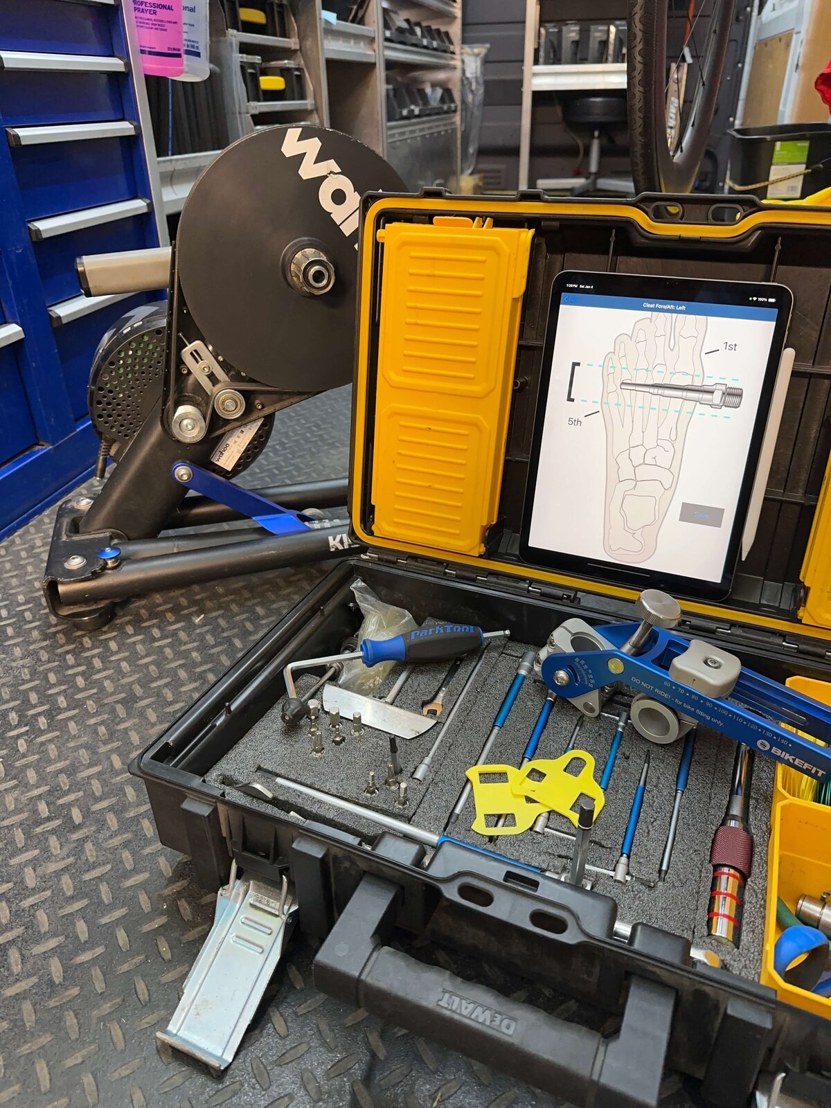 A bike repair station with tools in a black case, including wrenches and screwdrivers. A digital foot measurement device screen shows foot measurements. A Wahoo indoor bike trainer is visible in the background on a textured floor
