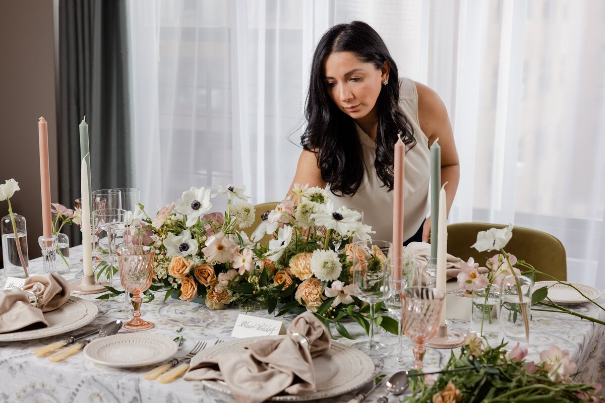wedding planner styling an elegant reception table with white florals, pastel candles, and delicate tableware in a bright, modern venue