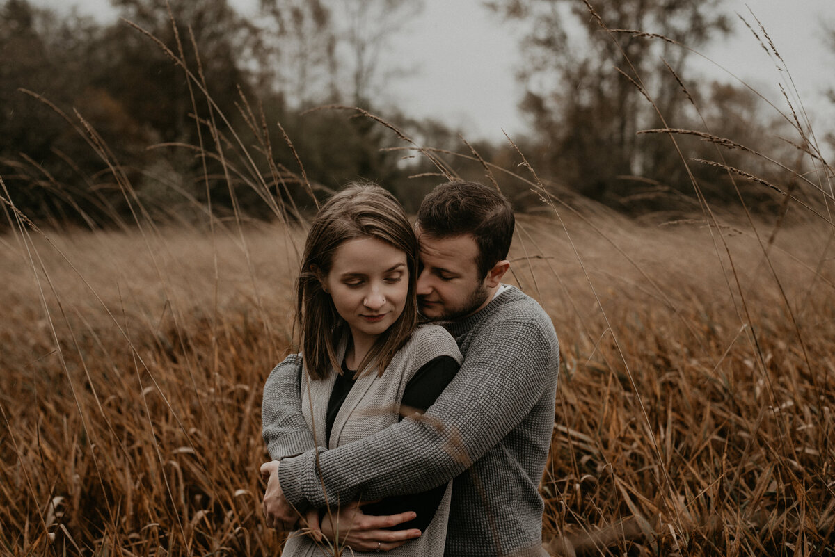 couple standing in the rain and wind in a field at campbell valley park. The guy is holding the girl from behind and they are nuzzled into each other intimately.
