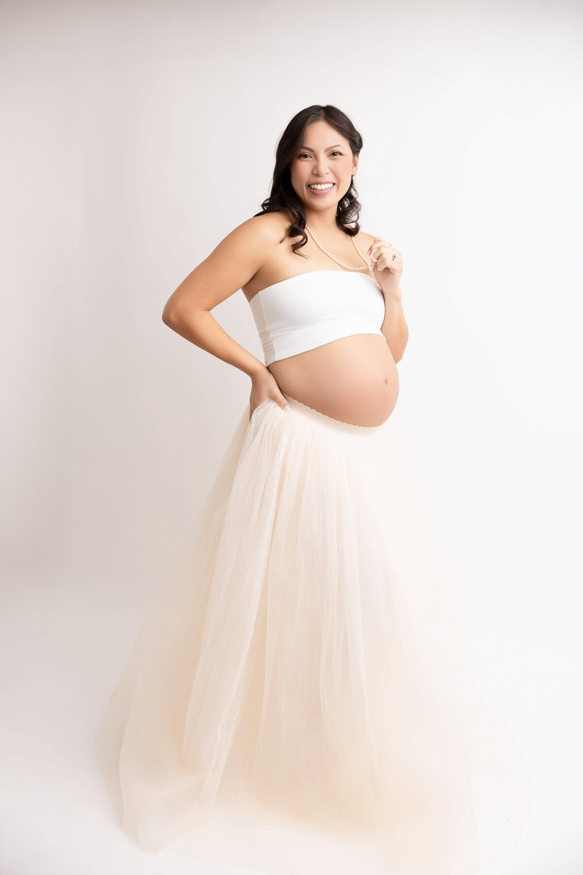 Smiling pregnant mom wearing a white tube top and pink skirt and posed against a white backdrop.