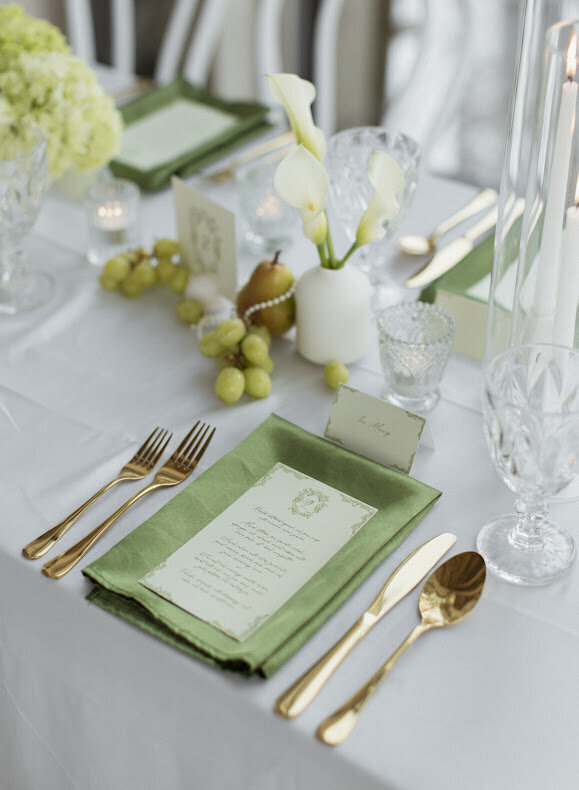 A close-up of a rococo-themed guest table at a National Arts Centre wedding, featuring collection of pears, pearls, bud vases and candles down the centre of the table. The table is adorned with white satin linens, green satin napkins and gold cutlery. An elegant pale green menu with Rococo detailing sits on top of the napkin.