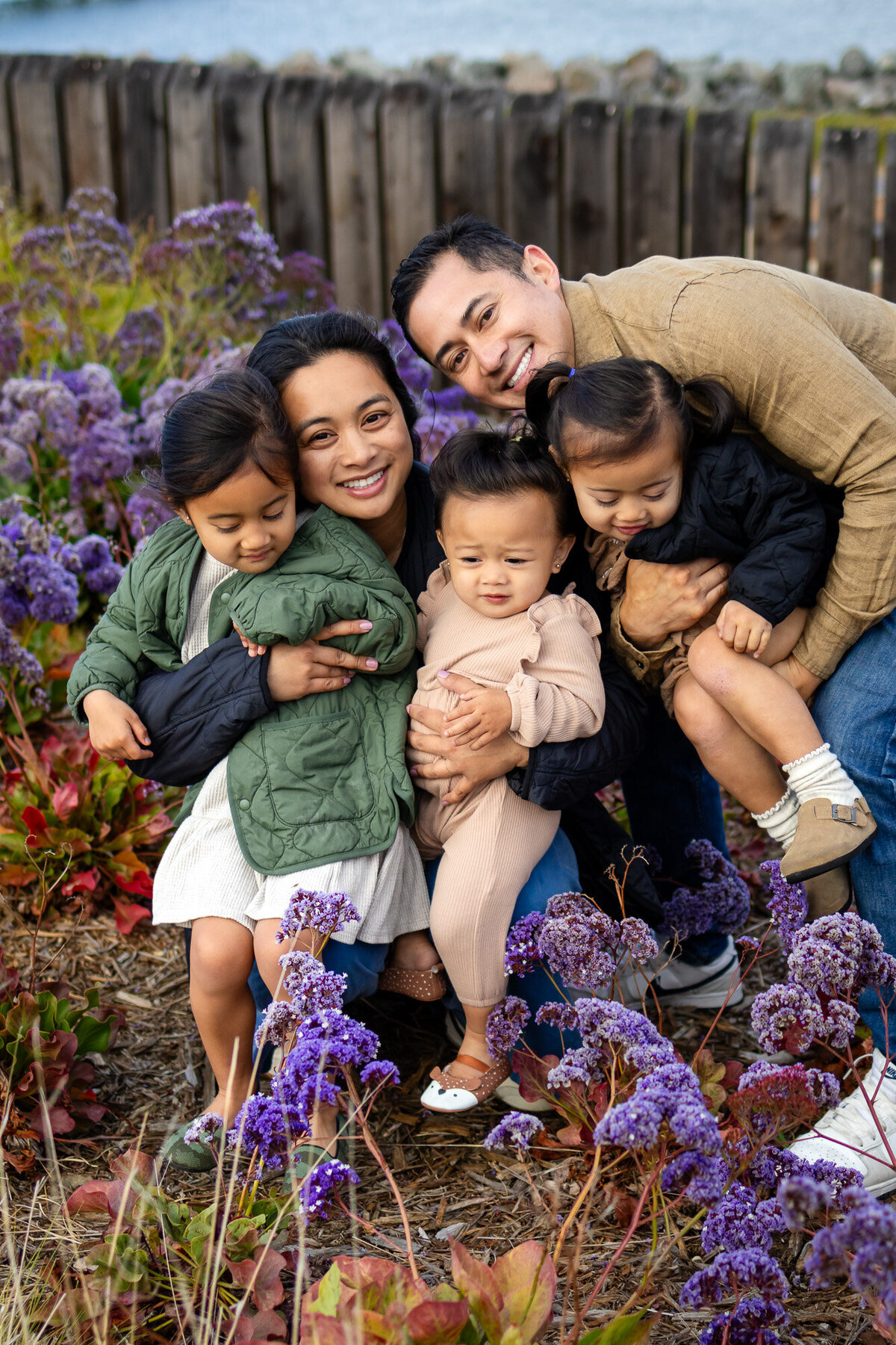 Bay Area Family Portfolio - Ellobelle Photography: Family of five cuddled together in front of blooming purple flowers near the water, with joyful smiles and natural connection.