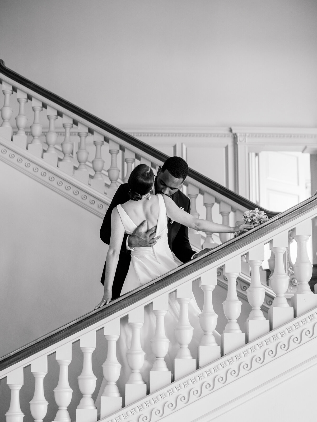 Black and white fine art wedding photograph of a romantic embrace on the grand staircase at Cairnwood Estate, captured by Philadelphia Fine Art Wedding Photographer Elizabeth Kane. This timeless image highlights classic East Coast wedding elegance, architectural beauty, and emotional storytelling.