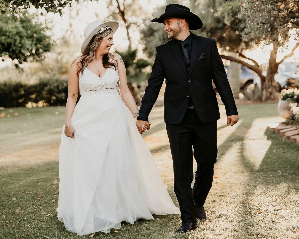 A groom wearing an all-black suit with boots and a cowboy hat holding hands with his bride.