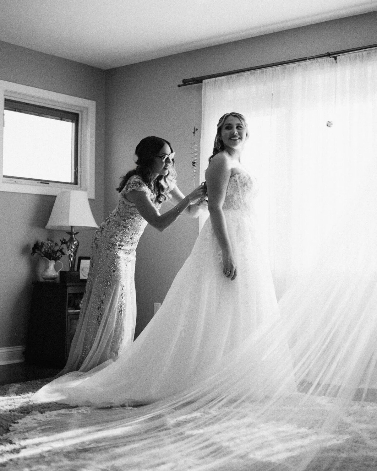 Two women in elegant dresses stand in a softly lit room; one helps fasten the back of the other’s wedding gown. Captured by a film photographer NJ, the bride smiles, framed by sheer curtains and sunlight streaming through the window.