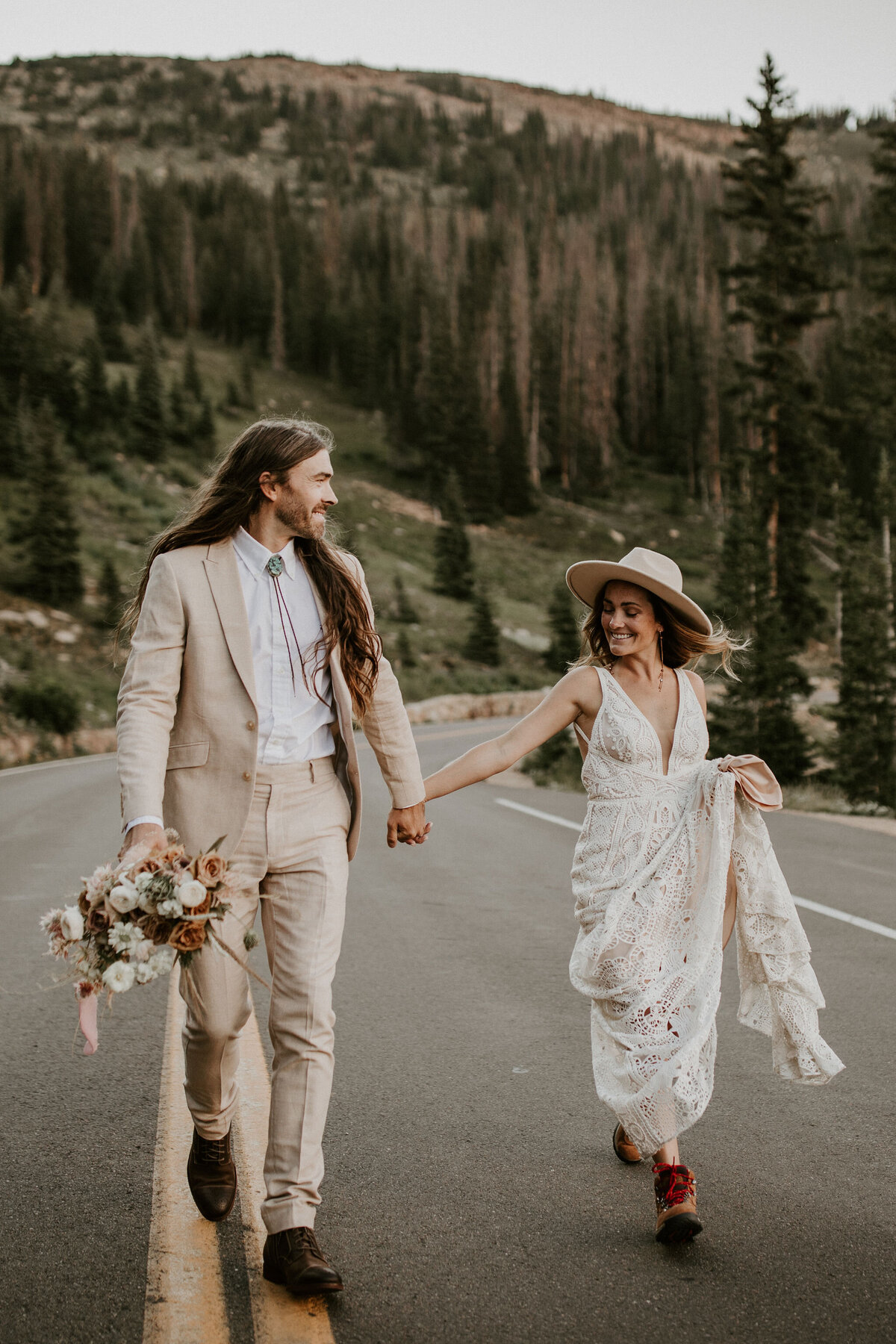 bride and groom wearing an ivory wedding gown and tuxedo hold hands while walking on a road in the mountains
