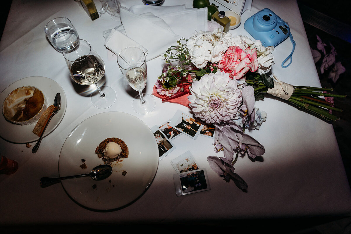 Late night photo of flowers, champagne, polaroid images and half-eaten wedding cake