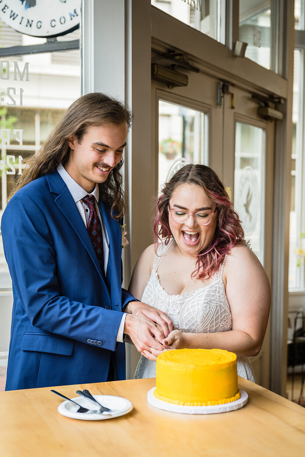 A couple holds onto a knife together to cut into a vibrant yellow cake on a long table inside of Olde Salem Brewery in Downtown Roanoke. The pair smile and laugh excitedly as they cut into their cake.