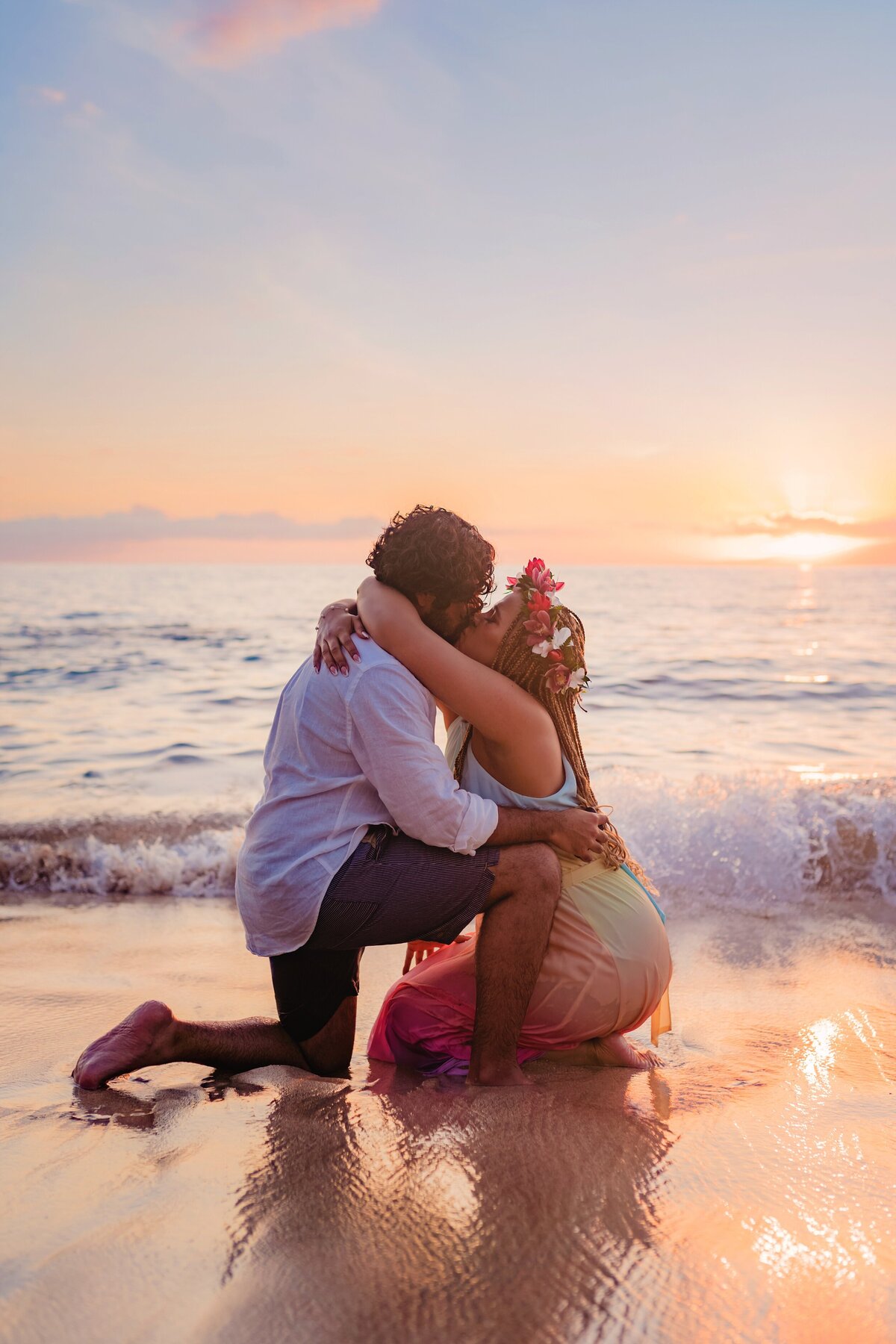 Woman kneeling down leans in to kiss her now fiance after he proposed on the beach in Maui