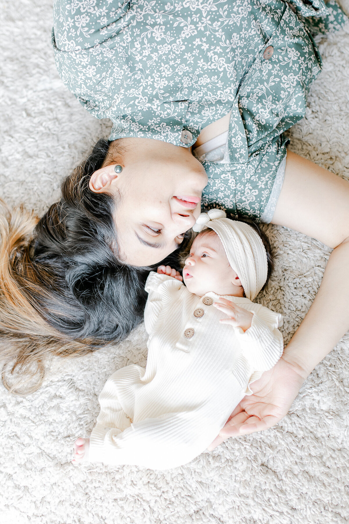 A tender mother-daughter moment – beautifully captured by a Birmingham newborn photographer. A new mom and her baby girl share a quiet connection, lying side by side on a soft, neutral rug. The warm, natural light enhances this sweet and intimate lifestyle newborn session at home.