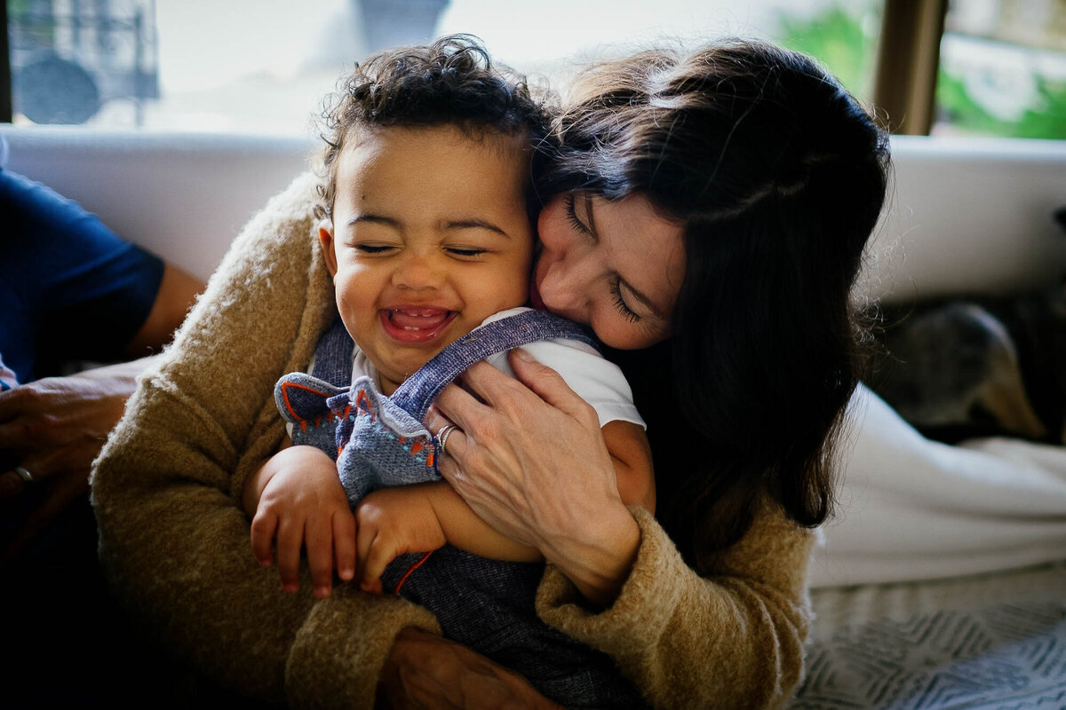 A mom squeezes her toddler son as he laughs joyfully during their at-home family photography session.