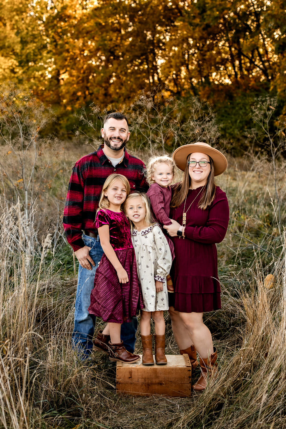 A Bloomington family photographer captures a family of five posing in a grassy field with autumn trees behind them. The adults, clad in dark, warm tones and one sporting a hat, smile alongside their three children—two in dresses and one in pants—for enchanting outdoor portraits.