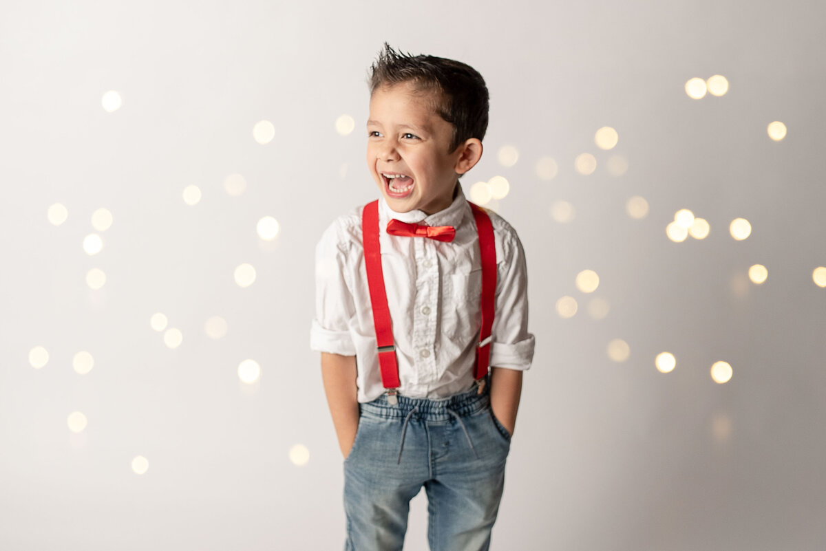 Toddler boy in portrait studio  wearing white button down shirt, jeans, red suspenders and red bow tie looking to the side and smiling really big. White backdrop and twinkle lights in the background.
