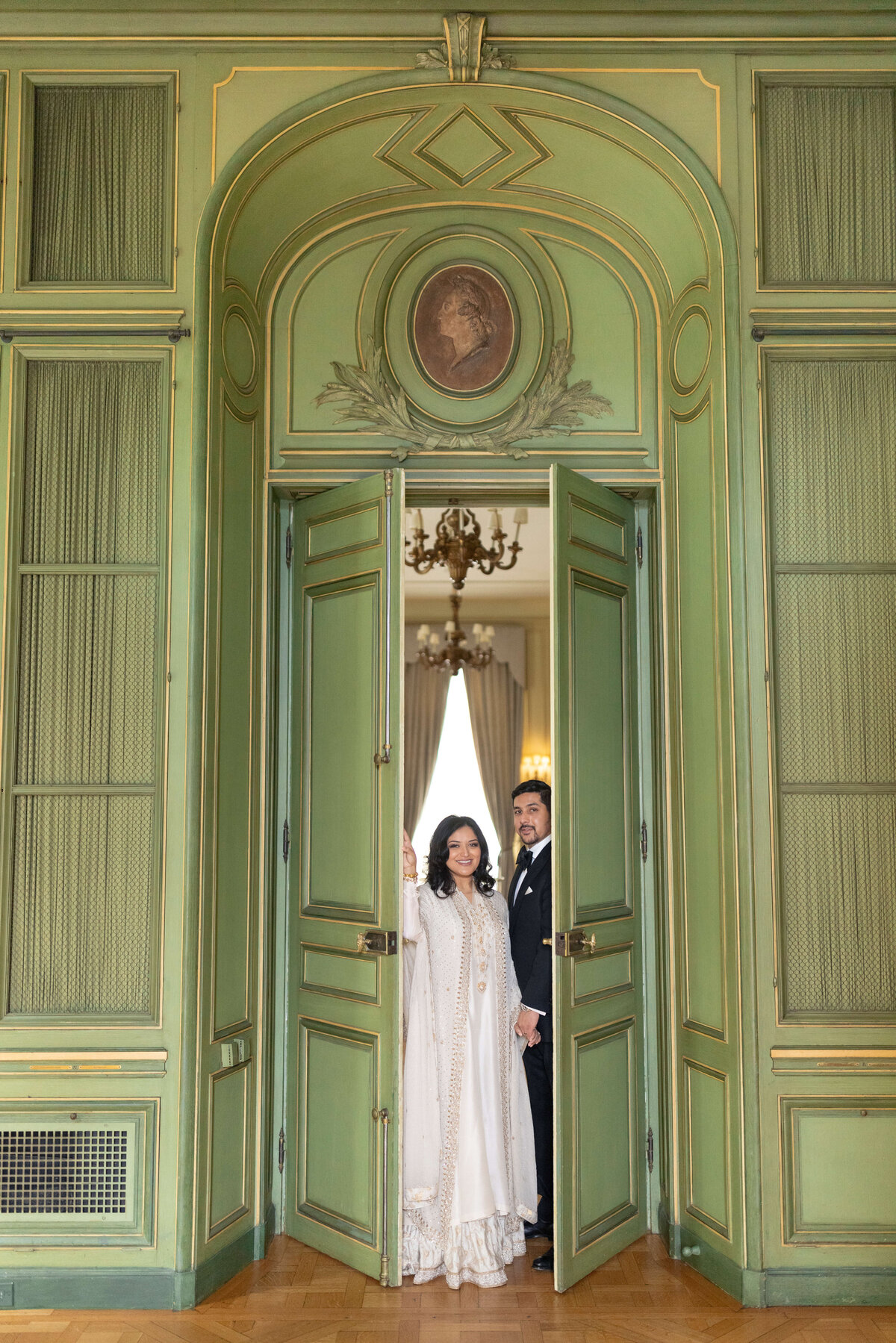 A couple stands smiling in an elegant room with green paneled doors. The woman wears a white dress, and the man wears a black suit. They are framed by the doorway, with a chandelier and curtains visible in the background.