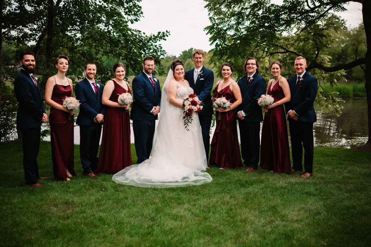 Newlyweds stand smiling with their wedding party by a pond
