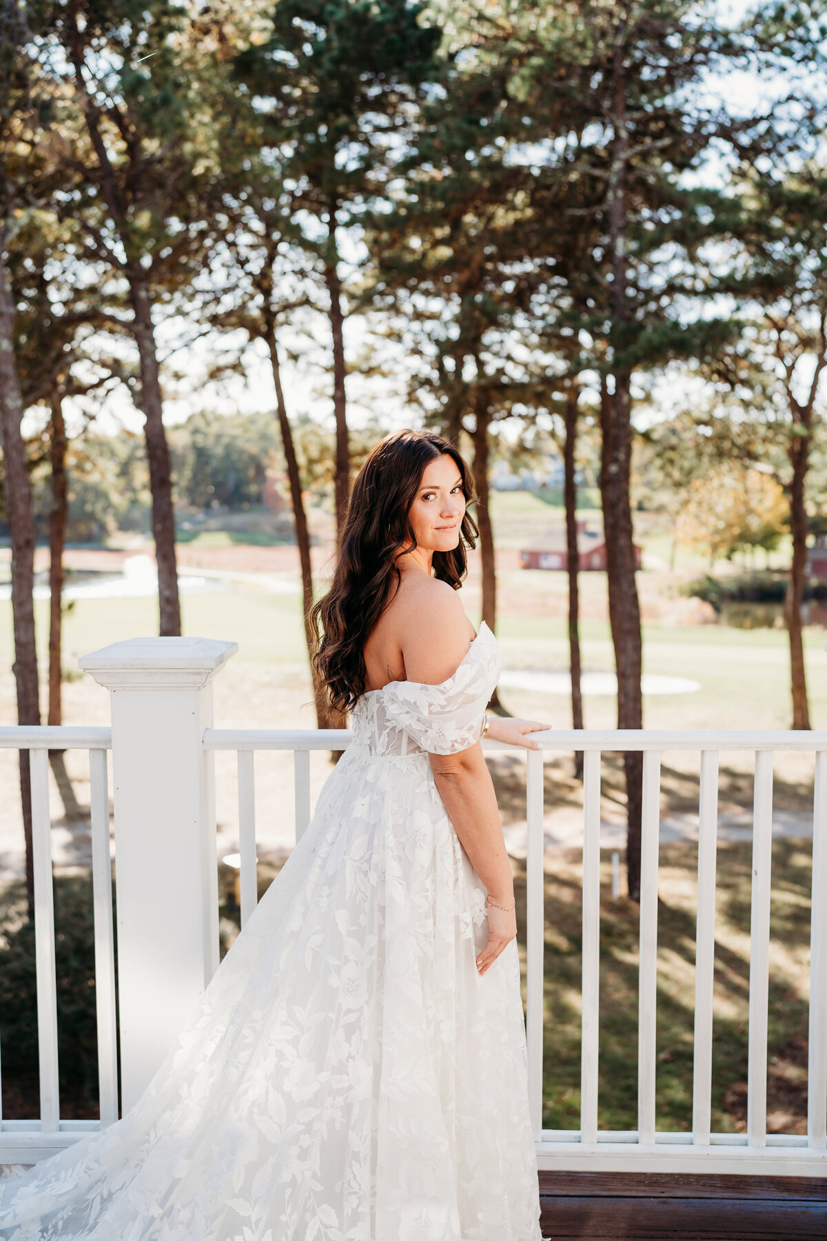Elegant bridal portrait featuring a stunning bride in an off-the-shoulder lace wedding gown, bathed in golden natural light. Captured on a scenic outdoor balcony with a breathtaking wooded backdrop, this image showcases timeless beauty and romantic bridal style. Perfect inspiration for outdoor weddings, fall weddings, and natural light wedding photography. Captured by Moll – Boston wedding photographer specializing in candid and artistic wedding photography.