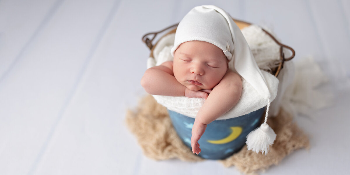 Newborn baby sleeping with arms folded over the edge of a hand-painted moon-themed bucket while wearing a white sleepy cap.