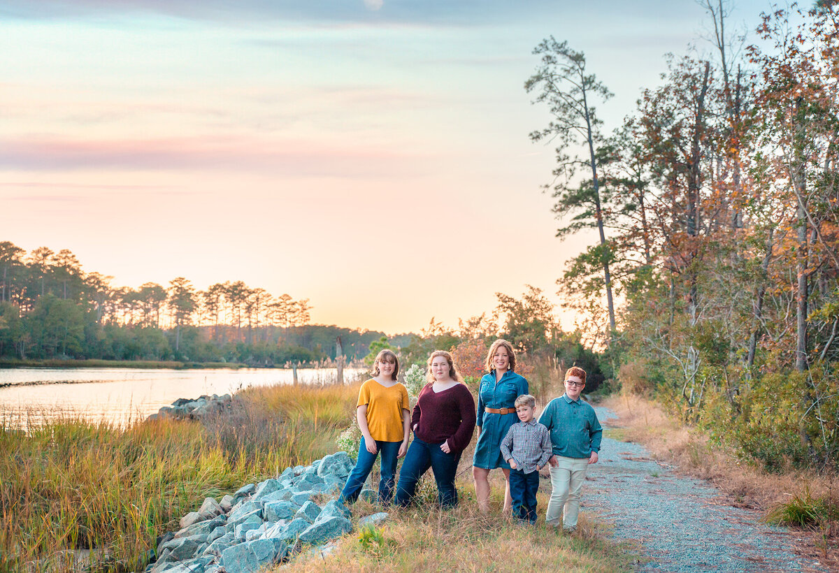 Hampton Roads family photos with parents and children posing on a gravel road next to the forest with the water in the distance as the sun sets creating a pink sky