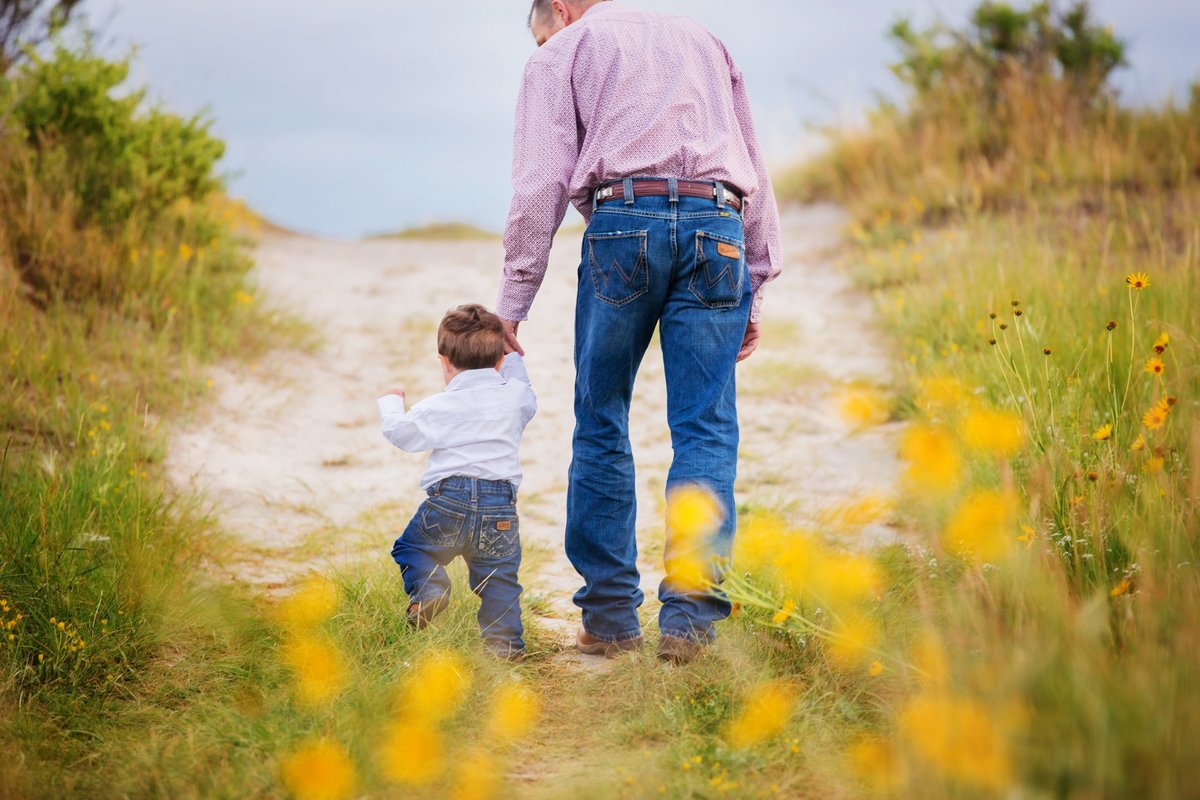 A toddler wearing wranglers holding his dad's hand while walking through yellow flowers in Laramie.