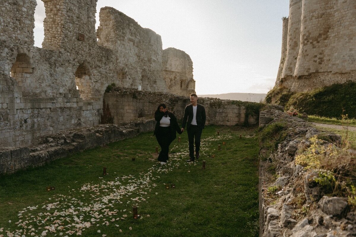 Couple main dans la main marchant sur un chemin de pétales dans des ruines médiévales. Séance engagement en Normandie, photographie par Kamescop.