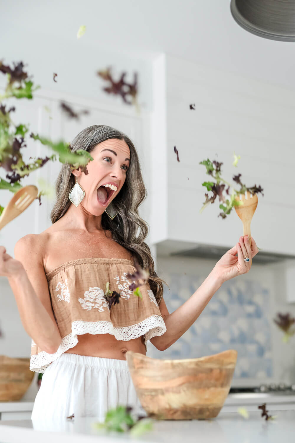 Woman in cropped embroidered top tossing leafy greens with wooden salad utensils mid-air.