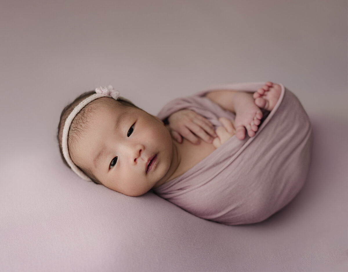 baby wrapped in pink swaddle with open eyes laying on pink backdrop