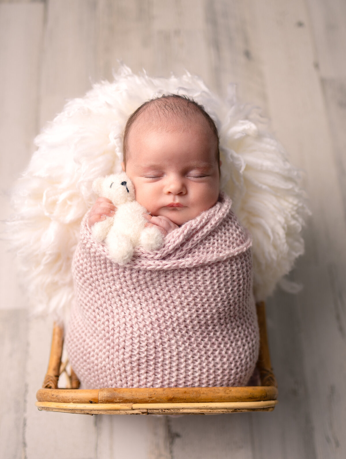 Newborn baby girl swaddled in a pink knit wrap holding a tiny white teddy bear during a professional studio session in NJ