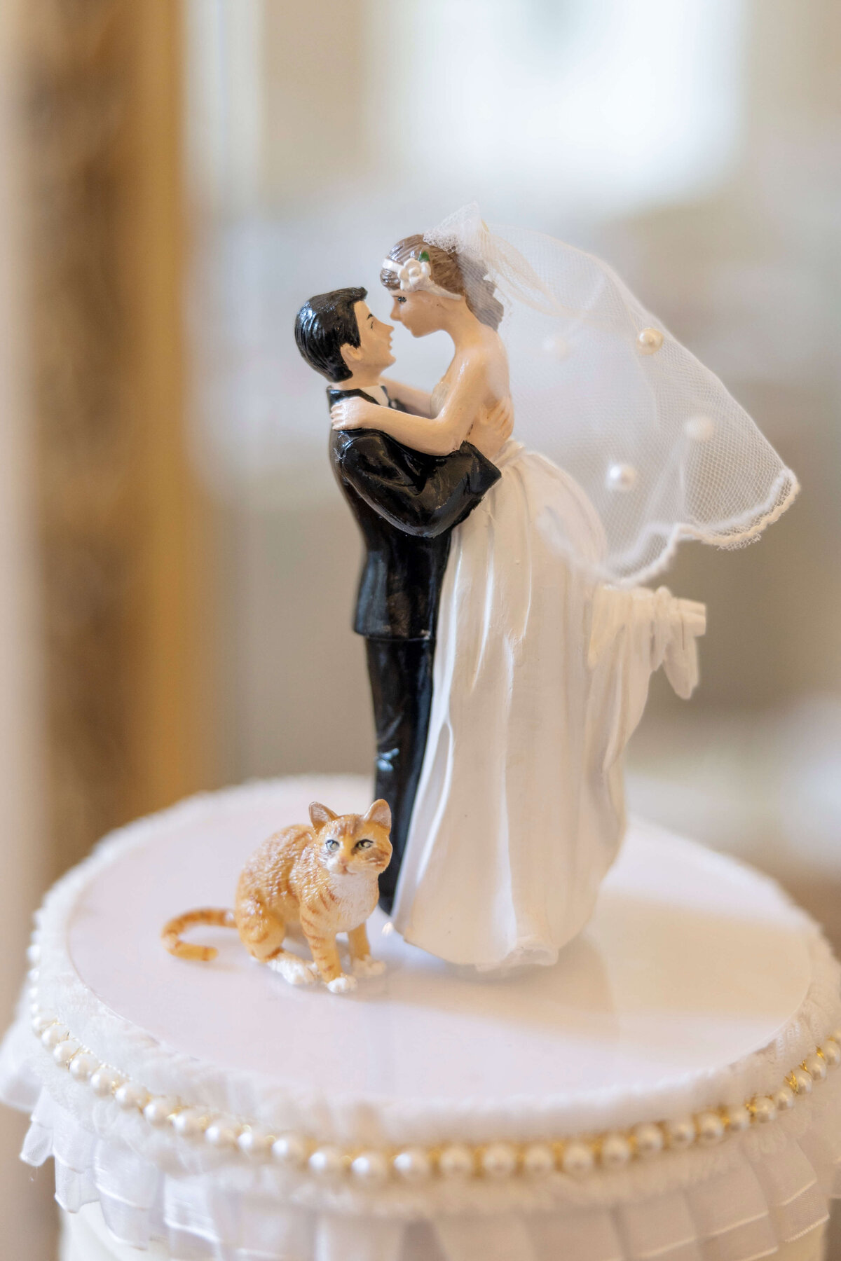 A wedding cake topper featuring a groom in a black tuxedo holding a bride in a white dress and veil. A small orange and white cat stands nearby on the cake, which is decorated with pearls along the edge.