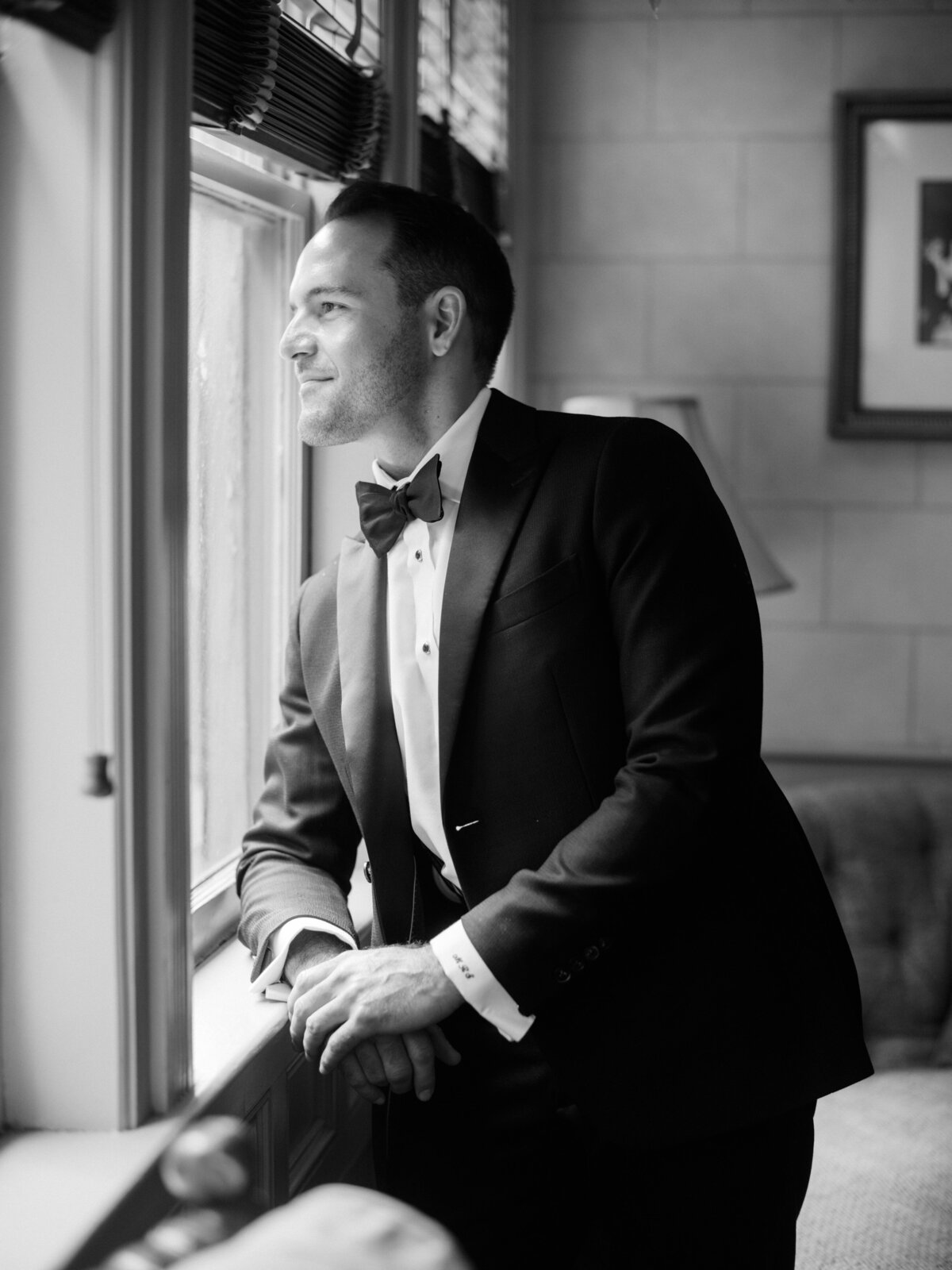 A dapper groom gazes thoughtfully out the window, reflecting on the moment before the ceremony while dressed in a formal tuxedo and bow tie, embodying elegance and anticipation.