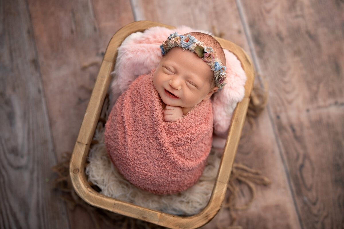 newborn baby girl wrapped in salmon colored knit wrap and wearing floral headband swaddled and posed inside rectangular wooden basket on wooden floor bacdrop