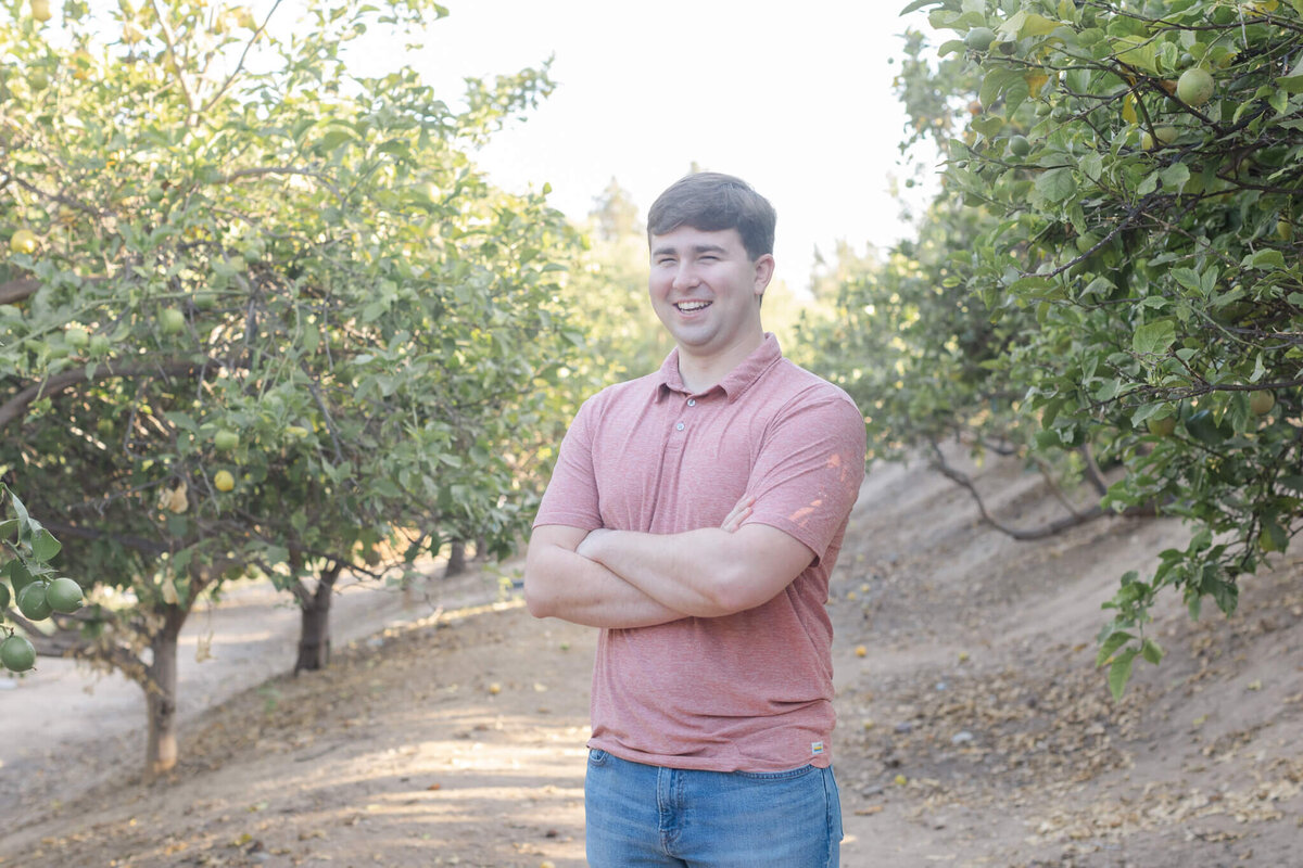 A young man with dirty blonde hair wearing blue jeans and a salmon shirt crossing his arms over his chest and smiling at the camera between two groves of green lemon trees.