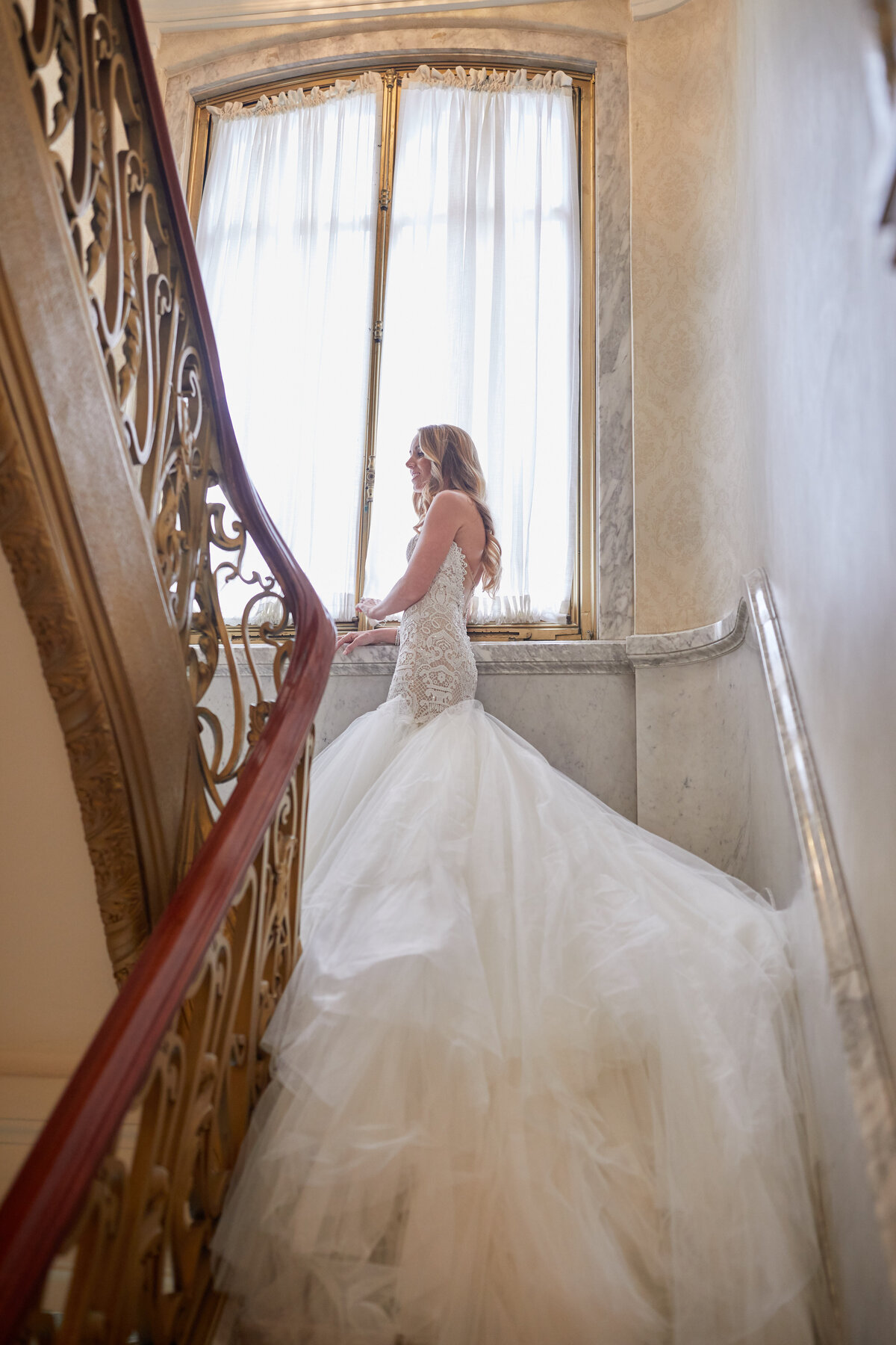 This stunning photo was taken at The New York Public Library in New York City by talented photographer Christian Oth. The scene captures a beautiful bride standing on an ornate staircase, wearing a lace and tulle wedding gown with a long train. As she glances down and delicately holds the railing, soft light streams in from a large window behind her, illuminating her dress and casting a gentle glow, creating a truly magical and elegant moment.   Additionally, East West Films provided videography services for this event, capturing the beauty of the bride's special day on film.