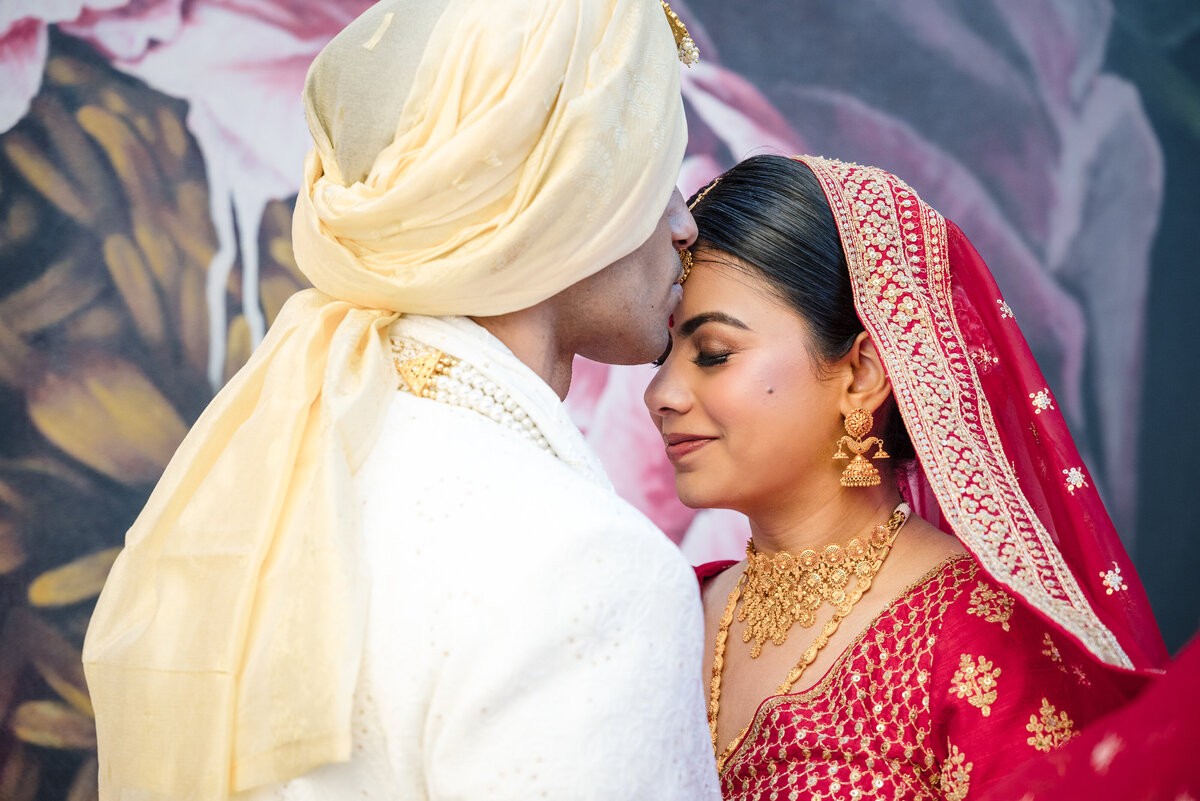 Indian wedding couple portrait at the Grand Bohemian Hotel Charlotte — groom in traditional ivory sherwani and turban kisses bride in red lehenga with gold jewelry. Captured by DeLong Photography, Charlotte Indian wedding photographers specializing in vibrant, cultural wedding photography