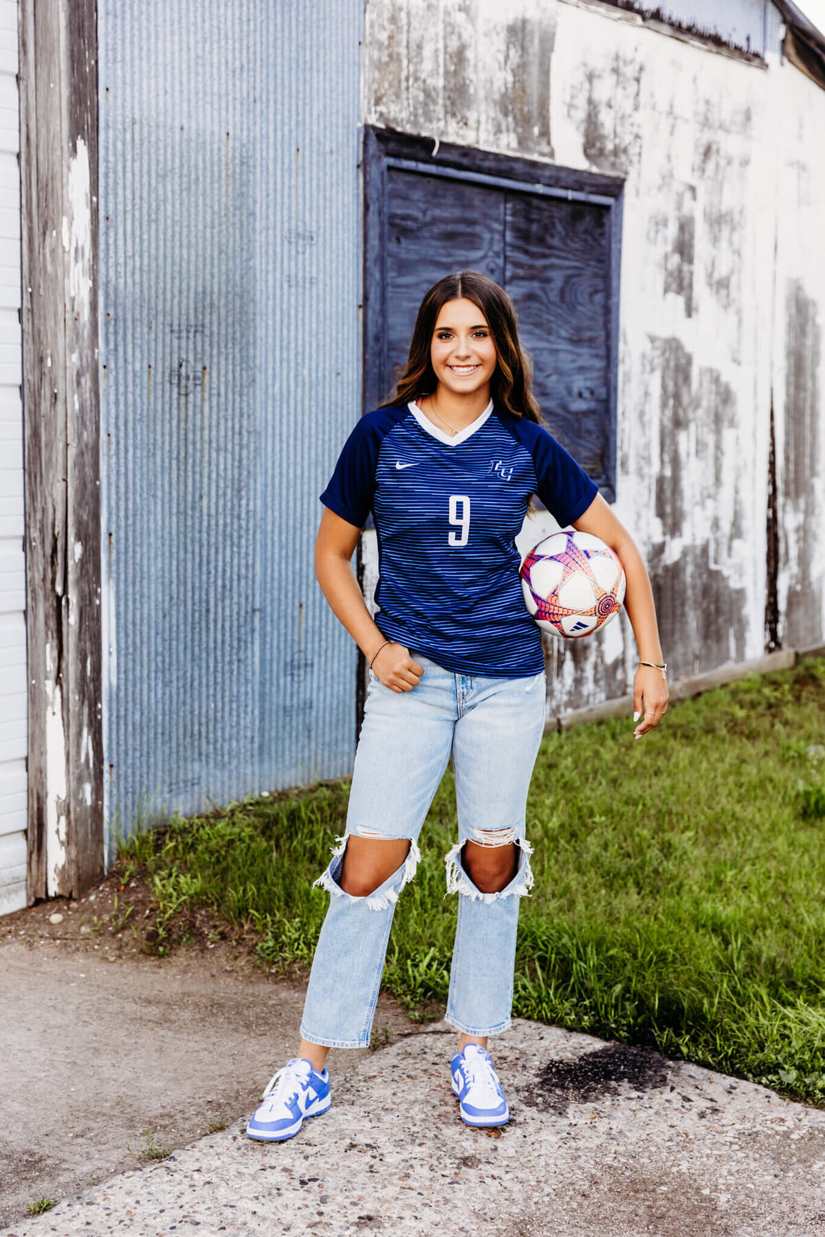 teen girl smiles as she poses with her soccer ball while wearing her soccer jersey and jeans 