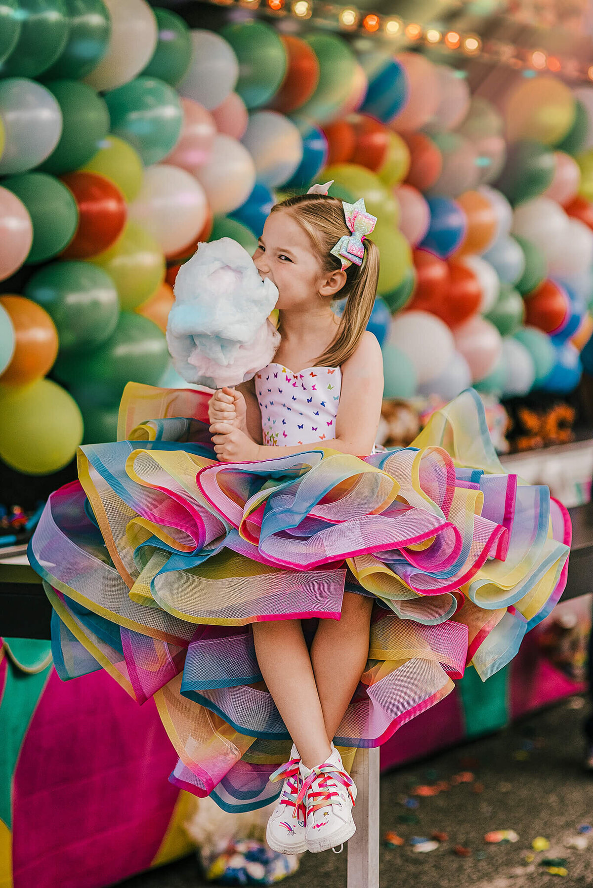 Girl in pigtails wearing a rainbow dress sitting in front o f balloons eating cotton candy at a carnival near Annapolis Maryland