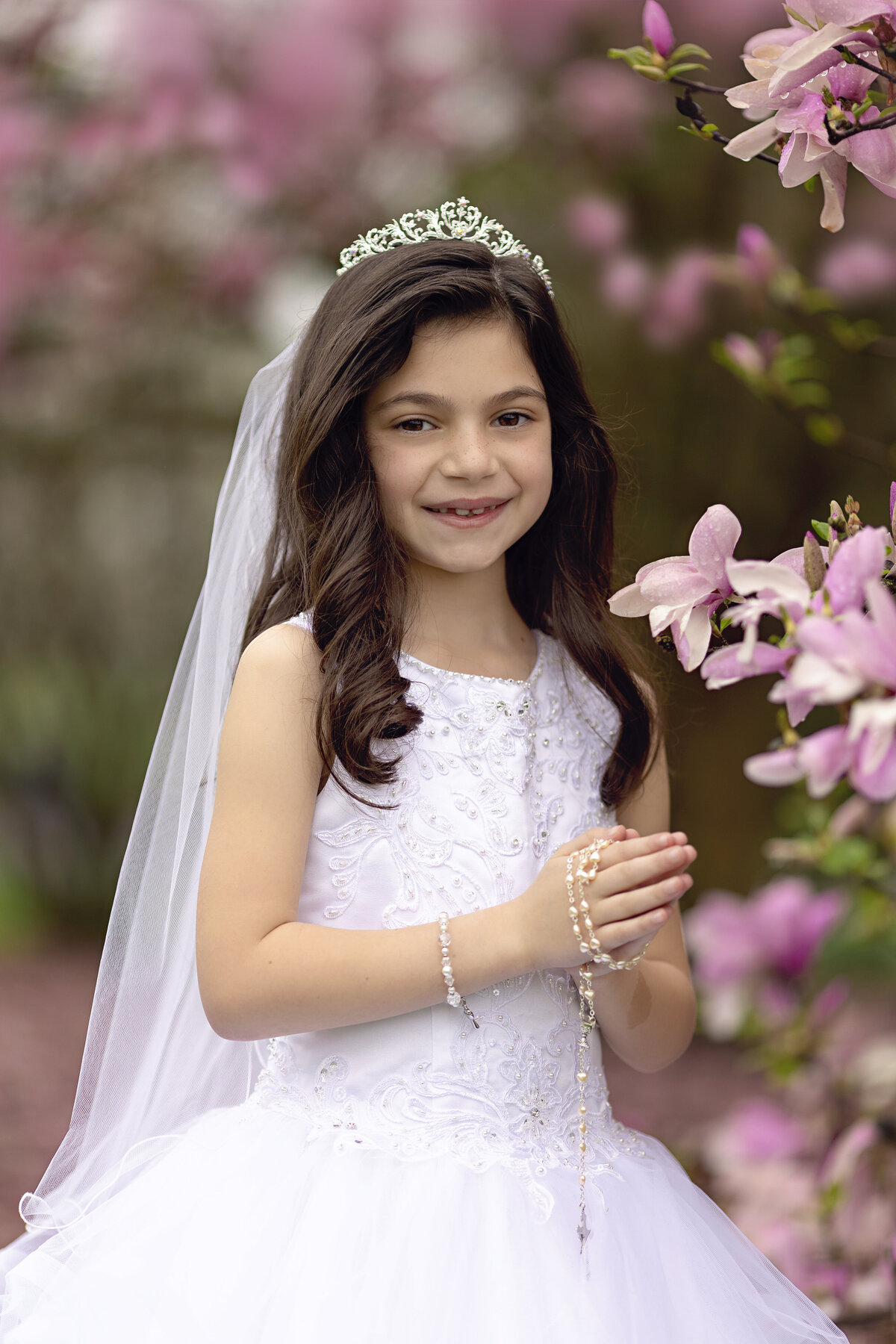 A young girl in a white lace dress prays with rosary beads with some pink flowers