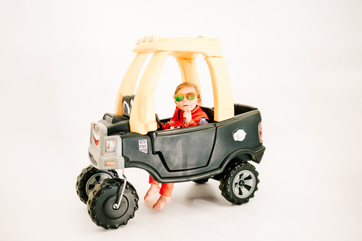 kid sitting in race car suit with glasses and car during his 1st birthday photography session