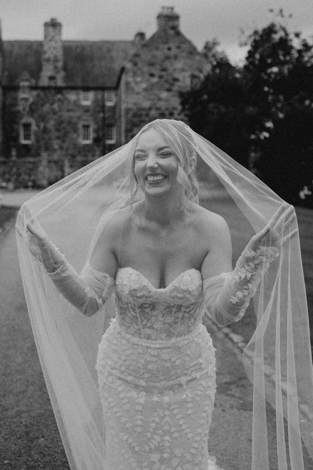 A bride playfully interacts with her veil. Barra Castle in Aberdeenshire is photographed behind her.