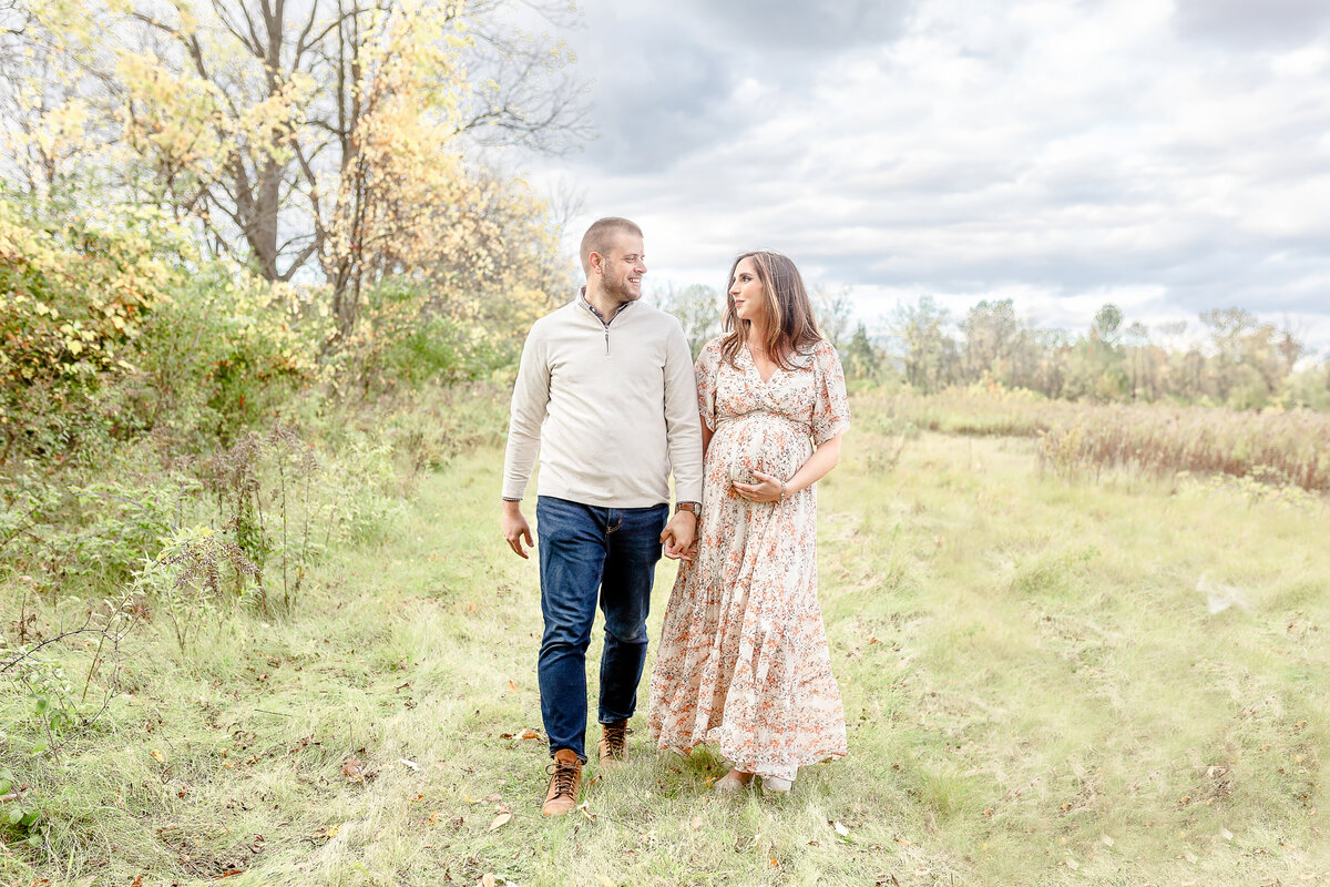 Couple walks holding hands while mom holds baby bump in Plymouth Michigan Newborn Photographer maternity session