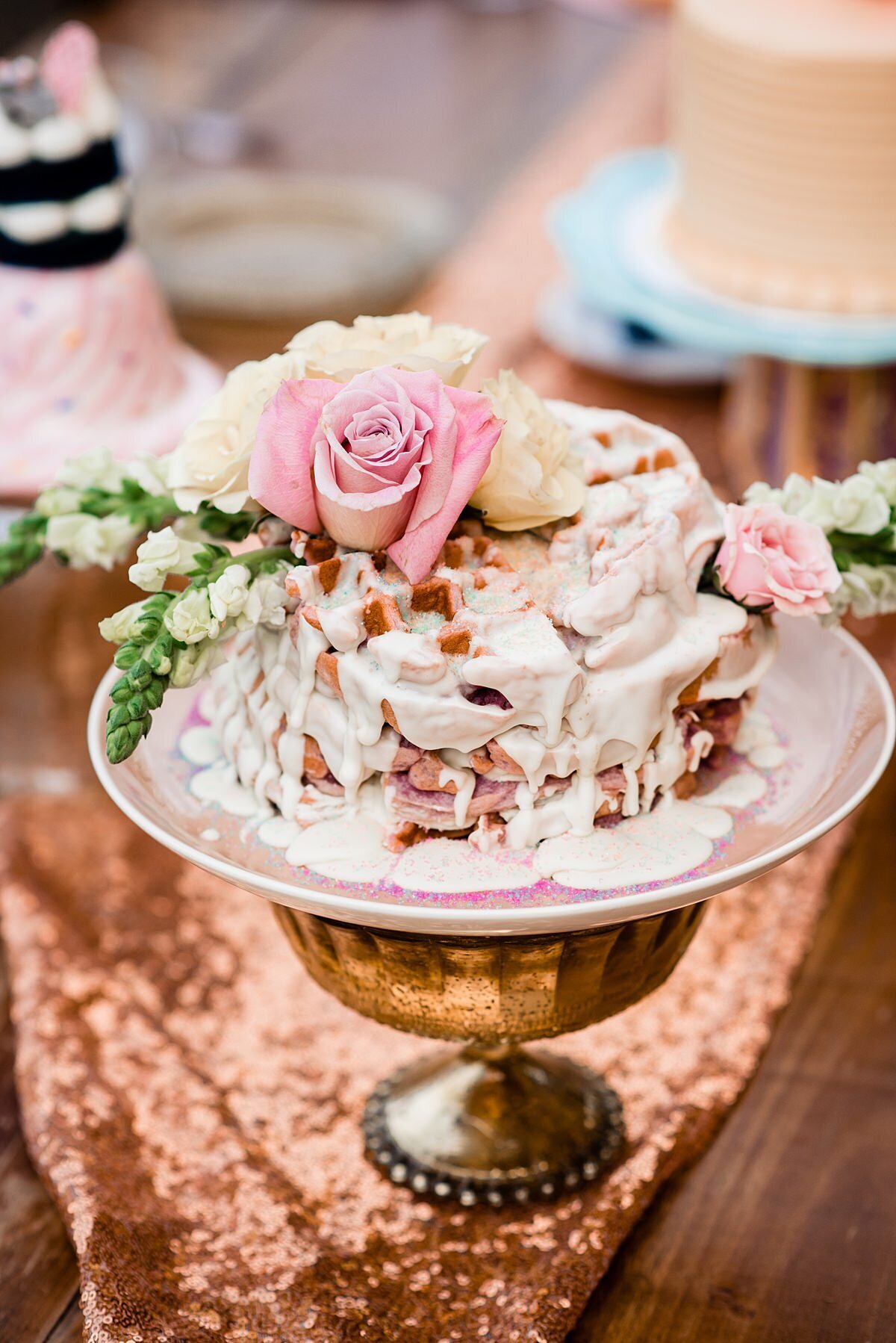 A purple waffle cake sitting on a pink plate drips with white icing as it sits on top of a silver footed stand. The waffles are decorated with pink roses, ivory roses and white stock. The farm table is decorated with a rose gold sequined table runner at Grace Valley Farm.