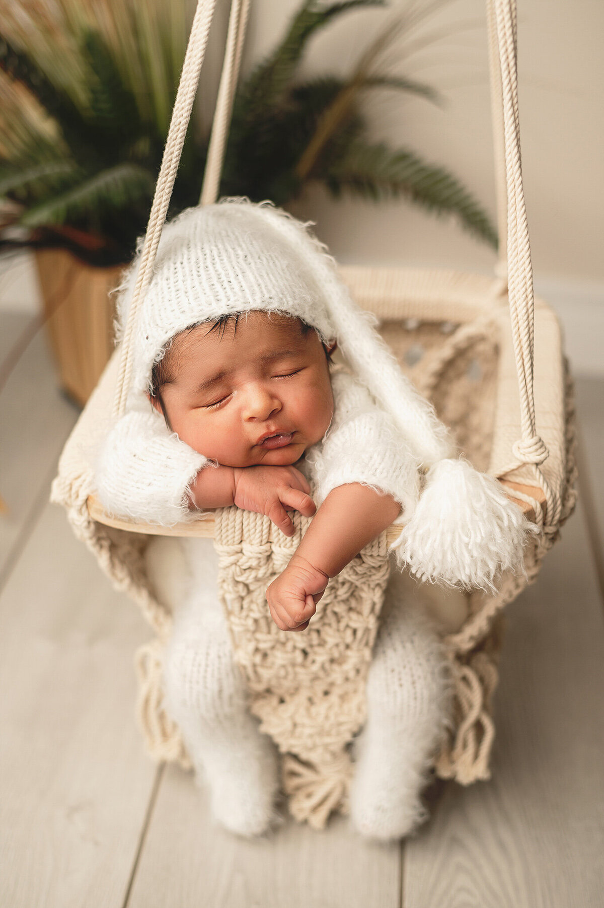 Infant baby boy in a white sleeper and hat sleeping in a macrame swing in his portrait studio session.
