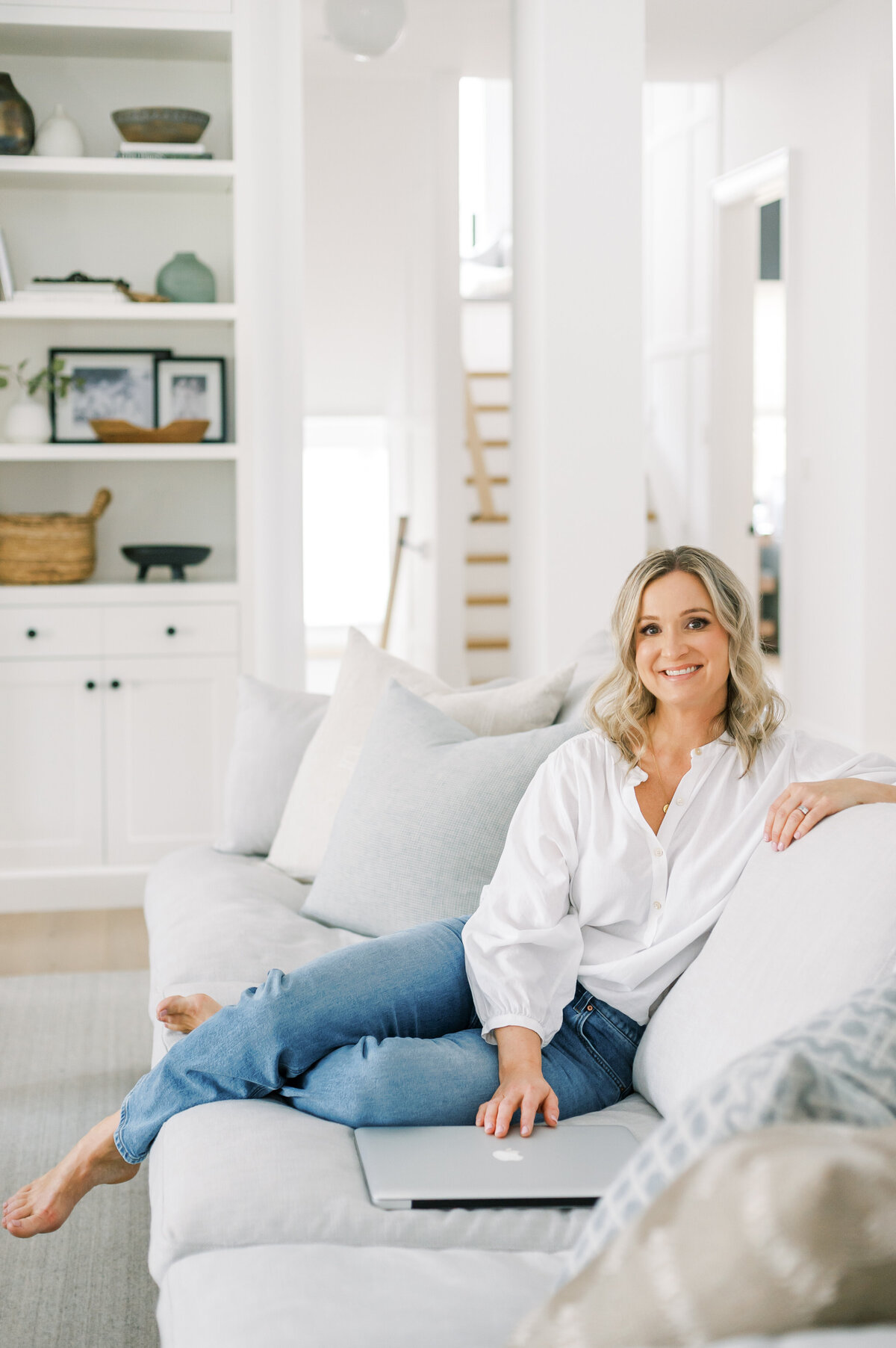Best Sarnia, ON interior designer and decorator, Jen Sottosanti, sits in her living room filled with light natural hues and natural wood tones. She is resting casually on the couch and smiling at the camera.