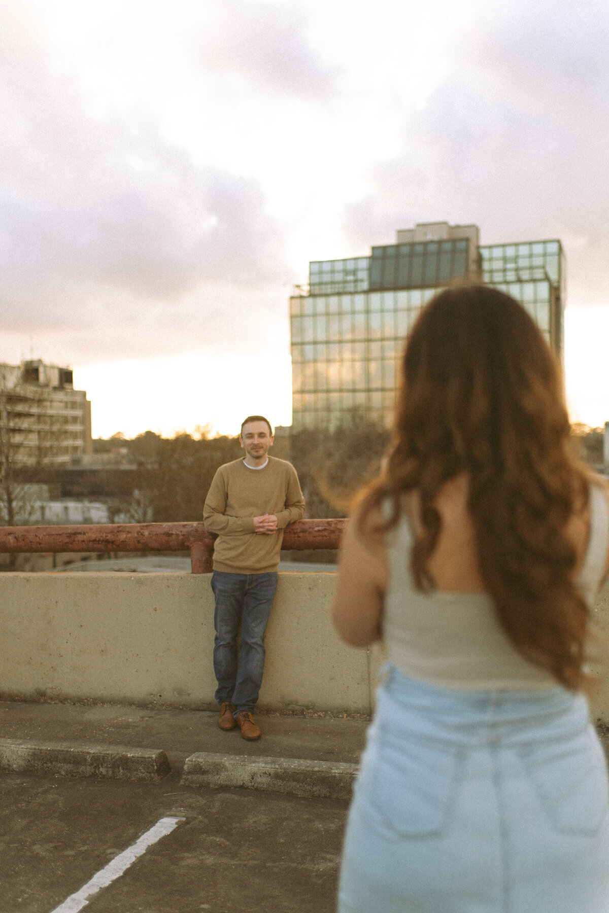 a woman taking a photo of her husband on top of a parking garage during golden hour. the city is their backdrop.