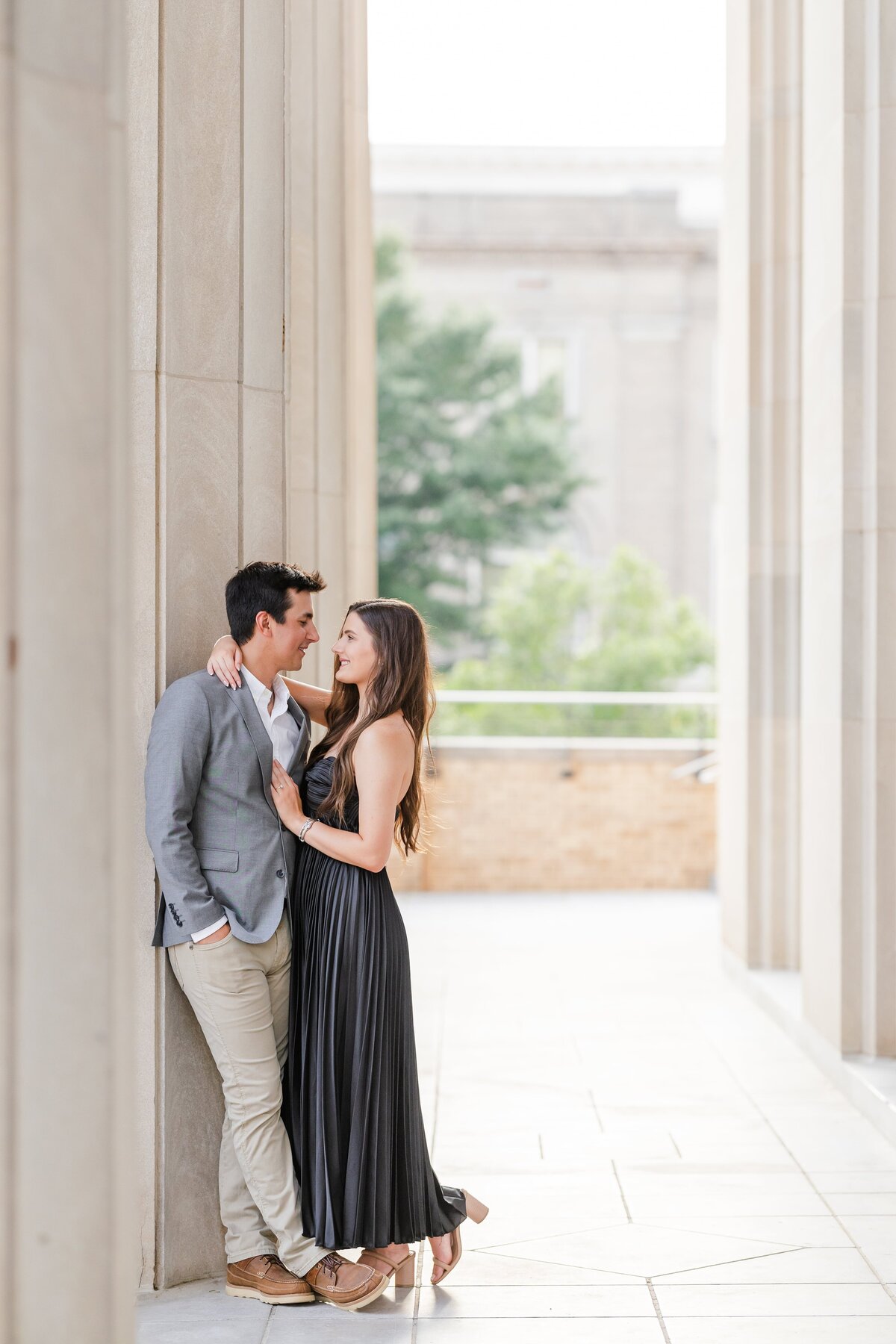 A happy engaged couple leans on a column hugging in a black dress and grey sports coat