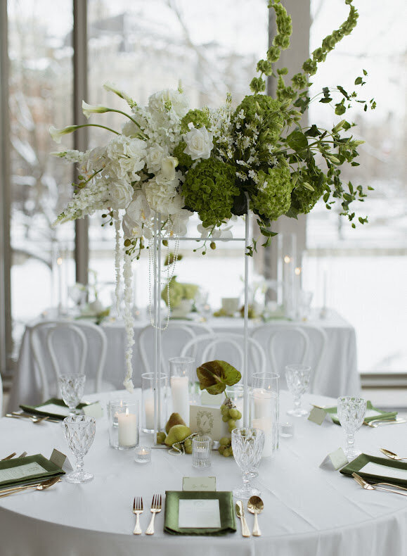 A close-up of a rococo-themed guest table at a National Arts Centre wedding, featuring a tall romantic colour blocked centrepiece made of white and bright green florals and a collection of pears, pearls, bud vases and candles at the base. The table is adorned with white satin linens, green satin napkins and gold cutlery.