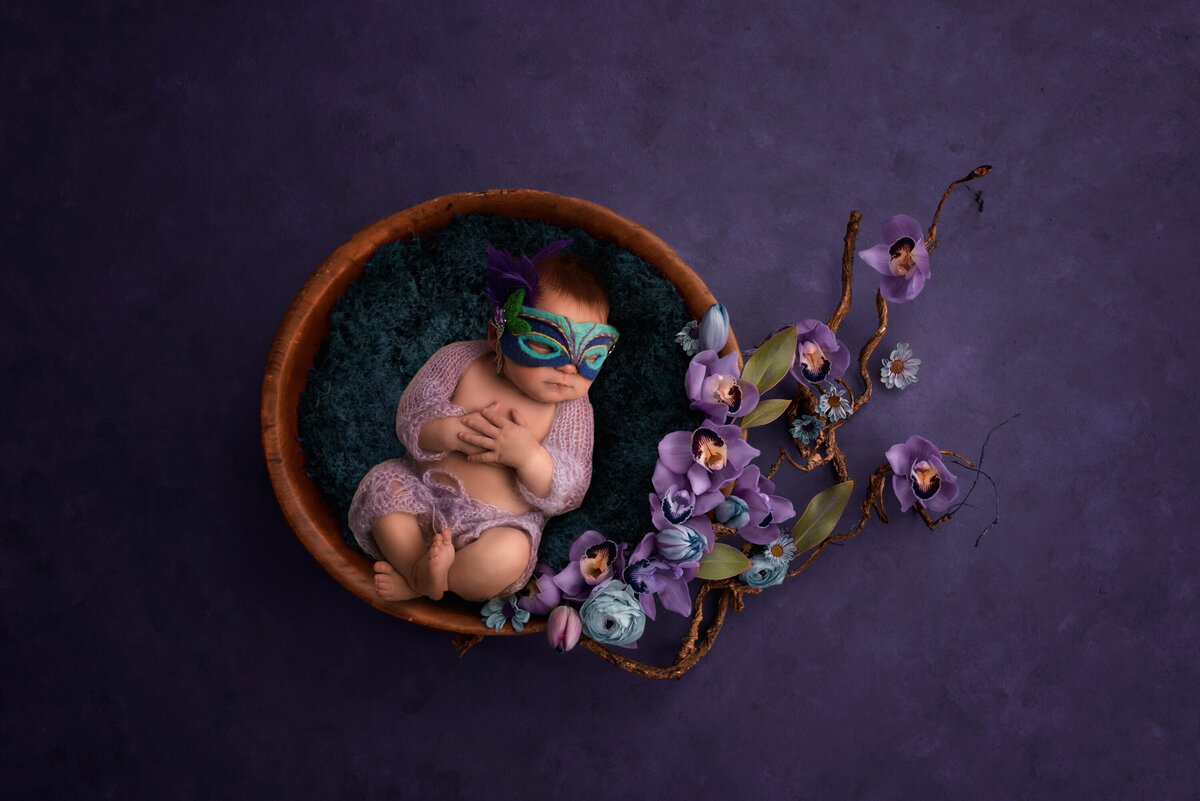 Newborn baby girl in lavender mohair pants and shrug with a Mardi Gras mask on.  She is in a wooden bowl on a purple backdrop with purple orchids.  Her hands are crossed over her stomach and her legs are crossed as well.