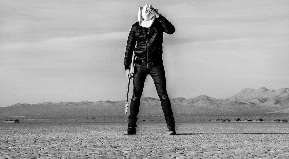 Country Musician black and white portrait Ben Klick standing in desert one hand holding onto white cowboy hat hes wearing shielding his face as he looks down other hand holding guitar El Mirage