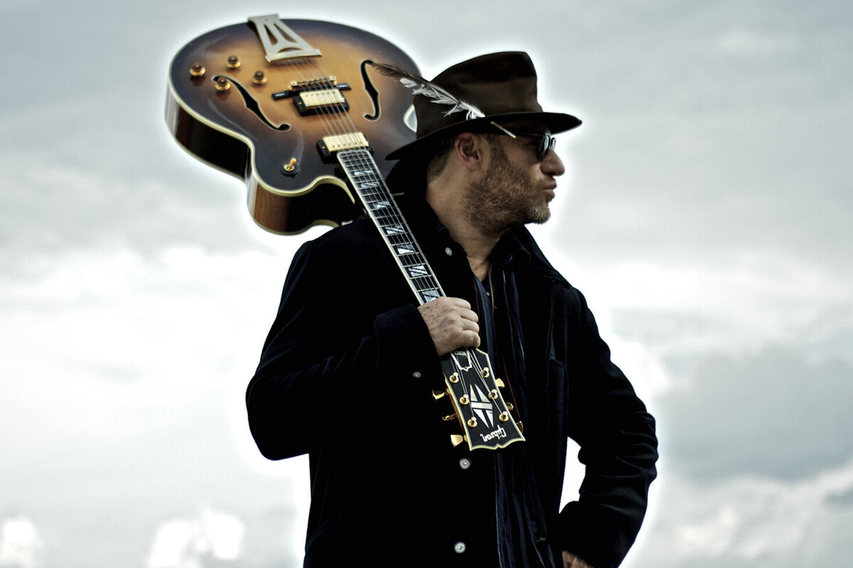 Music Artist Portrait Dave Sereny standing against cloudy sky holding guitar upright leaning against his shoulder looking off camera wearing hat and sunglasses Just Play Something Gallery